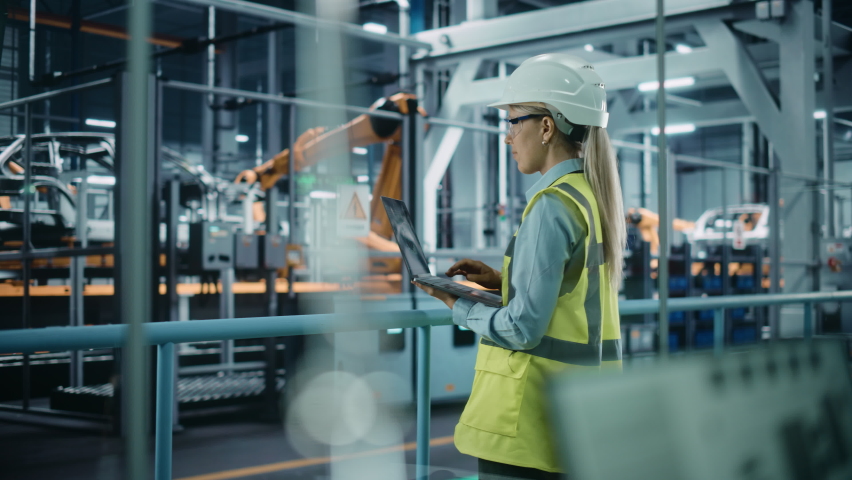Car Factory: Female Automotive Engineer Wearing Hard Hat, Standing, Using Laptop. Monitoring, Control, Equipment Production. Automated Robot Arm Assembly Line Manufacturing Electric Vehicles. Medium | Shutterstock HD Video #1077627434
