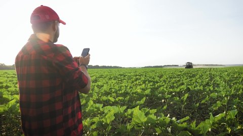 Farmer with tablet in front of tractor in field. Bearded agronomist wearing a cap uses a specialized app on a digital tablet PC on the background of working tractor in the field.