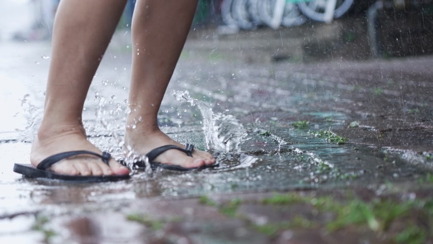 Low angle shot feet in flip flops stomping on water puddle on rainy day caused by monsoon, enjoys jumping and splashing water droplets around paving walkway, slow motion, tropical climate changes Royalty-Free Stock Footage #1077630878