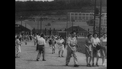 1940s: People walk up hill from buildings. People in front of Post Office. Family walks along strip mall. Man points to newspaper on wall. Domed building. Man works at panel. Man with equipment.