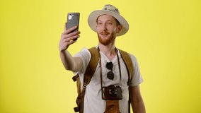 Happy Young Smiling Man With Tourist Backpack, Hat And Camera Talking With Friend Via Video Call On Smartphone On The Yellow Background. The Tourist Goes On Vacation, Traveling Concept People