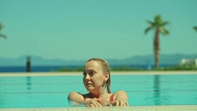 A young blonde woman in a swimsuit at the pool. Young woman in swimsuit cooling off with lemon juice or soda by the pool, enjoying her vacation. Slow motion video.