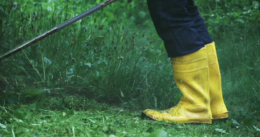 Competent gardener using electric hands mower for trimming high grass around house. Man wearing yellow rubber boots while sitting loan during summer time.
