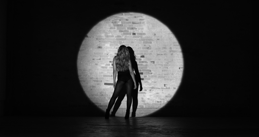 Beautiful young woman dancing seductively in brassier, black panties, and boot heels in the studio on a brick background with a focused stage light directed towards her, on a black and white video.