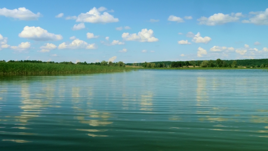 Panoramic view of blue sky with white clouds and nice view of river coast with green reeds and trees.
