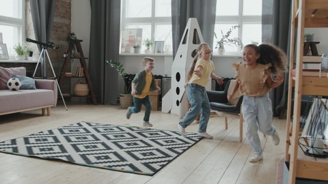 Handheld slowmo shot of happy 8-year-old girl with blindfold running and playing tag with group of little friends in cozy living room at home