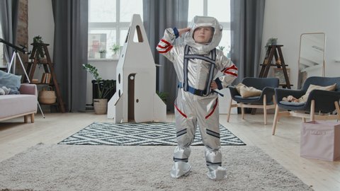 Portrait shot of cute 6-year-old boy in spacesuit posing for camera in living room at home and saluting. Cardboard rocket ship standing in background