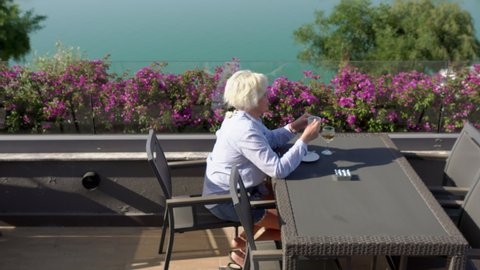 Blond woman enjoying a cup of coffee on an outdoor terrace overlooking a calm river viewed across tropical bougainvillea