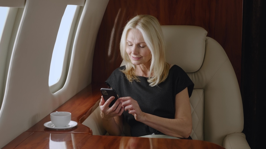 Beautiful smiling mature business woman using smartphone while travels on private jet plane. Rich senior woman using mobile phone and enjoying luxury journey in first business class airlines Royalty-Free Stock Footage #1077671423