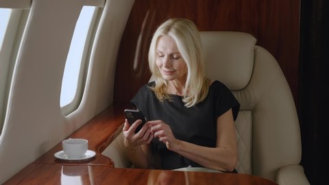 Beautiful smiling mature business woman using smartphone while travels on private jet plane. Rich senior woman using mobile phone and enjoying luxury journey in first business class airlines