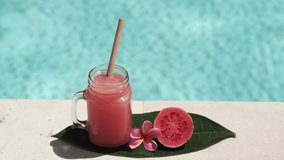 Video footage of glass mason jar with pink guava juice, bamboo straw, half of fresh pink guava, tropical flower frangipani and bubbling blue swimming pool on background.