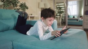 Curious Child Little school Boy Using Digital Tablet Technology Device Lying On Sofa Alone. Small Boy Hold Pad Computer Surfing Internet Play Game At Home. Children Tech Addiction Concept