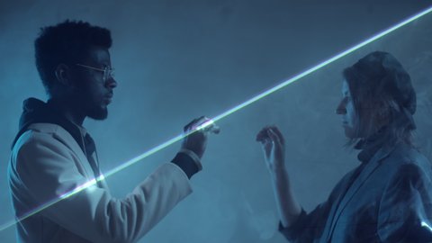 Side view of young black man and Caucasian woman standing in dark studio separated by flat sheet of laser light and holding hands