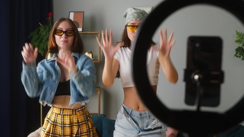 Two young sisters wearing sunglasses recording trend dance video on smartphone with lamp in living room. Female students, teenagers dancing for musical clip. Bloggers making music content for channel