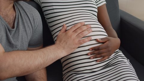 Close up of baby bump of pregnant black mother and caucasian father holding hand on belly. Interracial couple with pregnancy expecting child together on couch in living room.
