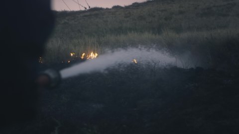 A firefighter holds a hose with water in his hands, extinguishes a burning flame. Natural objects are burning: grass, steppe, forests. Water from a hose is sprayed onto the fire. Close-up, slow motion