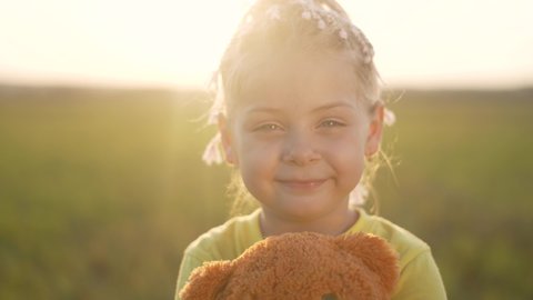 Happy little girl in park at sunset. Kid with toy teddy bear looks into camera. Girl smiles. Happy girl with friend teddy bear. Kid play with toy in park. Girl with friend in park at sunset.Baby smile