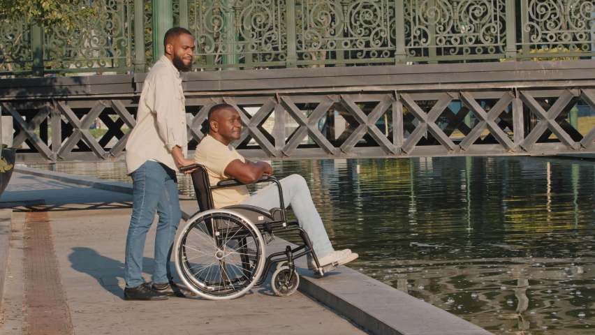 Young bearded African American guy, orderly, guardian or relative pushing wheelchair closer so that elderly disabled black man can admire water. Son accompanies sick father during walk in summer city | Shutterstock HD Video #1077701948