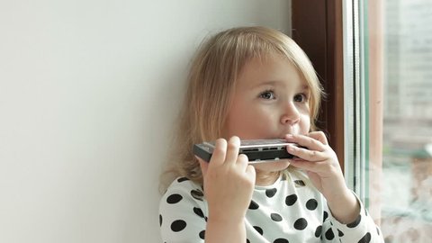 Little cute girl playing harmonica sitting by the window  