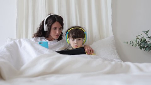 Mom help kid boy with educational lesson in digital tablet. Small boy lying in bed with mother hold touchscreen pc playing games, watching cartoons or doing homework for preschool education online
