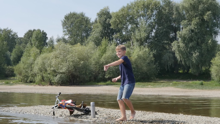 cute Caucasian teenager barefoot throws stones into the water while standing on the bank of the river. Leisure activities in nature. Slow motion.
