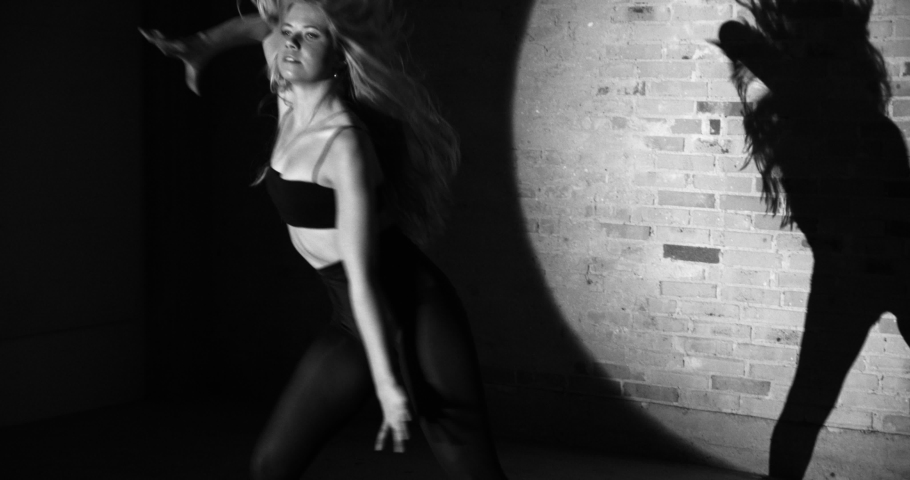 Steady shot of young beautiful dancer, prancing her body energetically, wearing brassier and high waist panties on brick wall with her silhouette shadow and focus light in background, black and white