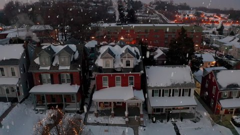 Snow falls at traditional two story American homes covered in winter snowfall, decorated with Christmas holiday lights. Aerial truck shot. Smoke from chimney.