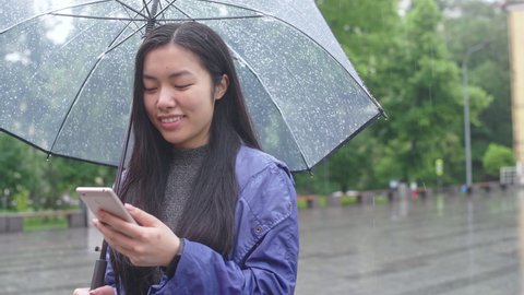 Happy asian woman scrolling social media on phone, waiting for friend under rain