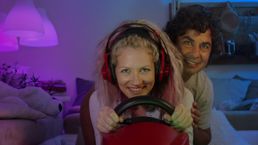 happy smiling couple at home in the evening, young wife uses the steering wheel playing a computer video game her husband looks scared and worried, woman drive car concept