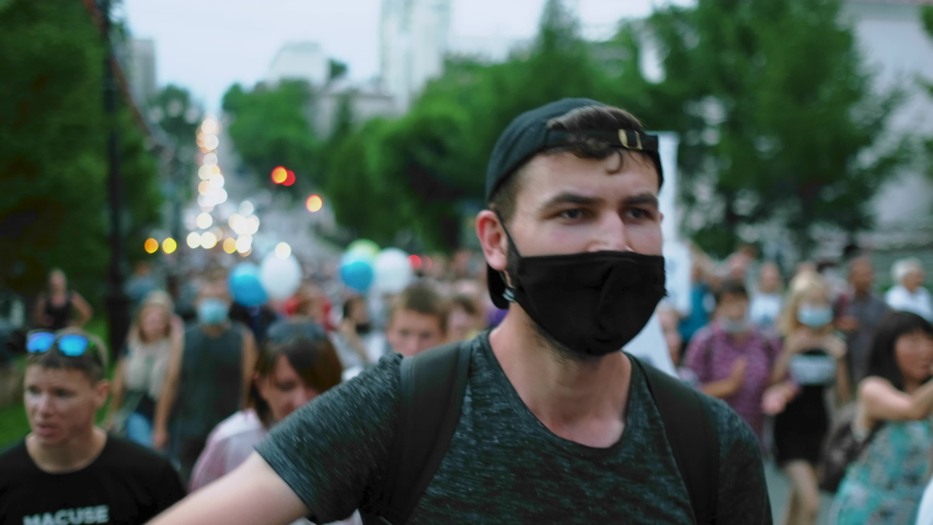 Masked face of male protester rebel in coronavirus restriction resistance crowd picket demonstration. Political opposition activist guy in covid-19 facemask waves arm fist. City riot, rally revolt. Royalty-Free Stock Footage #1077725351