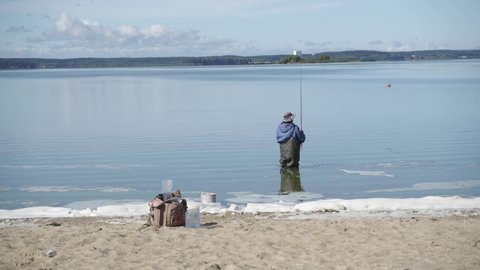 a man in warm blue and green clothes and a hat went into a lake or river to fish on a telescopic spinning rod near a sandy beach with foam, snow, ice and trees against a blue sky with clouds.
