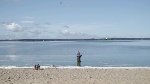 a man in warm blue and green clothes and a hat went into a lake or river to fish on a telescopic spinning rod near a sandy beach with foam, snow, ice and trees against a blue sky with clouds.