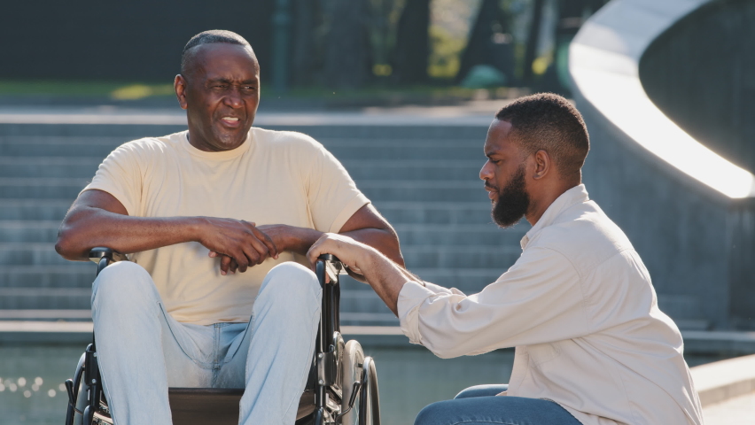Young African American guy squatting down talking to elderly father in wheelchair. Healthy black man friendly with disabled person. Patient care, equal opportunity, inclusive environment concept | Shutterstock HD Video #1077734465