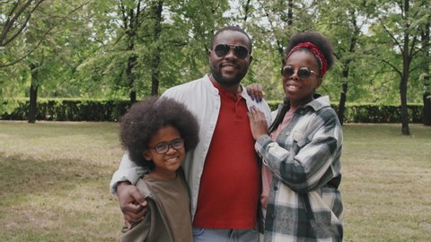 Medium slowmo portrait of modern African American family of three wearing casual clothes and sunglasses smiling to camera standing outdoors in park on warm summer day