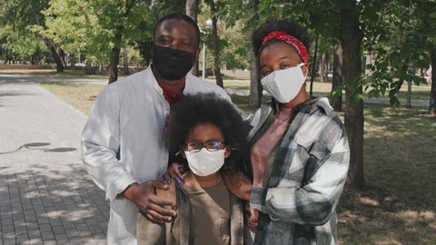 Medium slowmo portrait of happy African-American family of three wearing face masks posing for camera standing outdoors in park in summer. Parents hugging their cute son