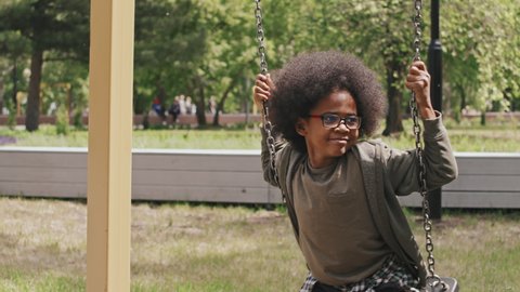 Slowmo shot of cheerful African-American boy swinging on chain swing outdoors on warm sunny day