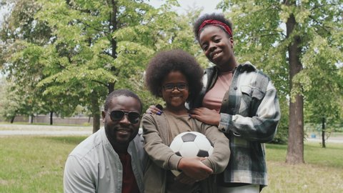 Low-angle medium slowmo portrait of happy African-American family of three posing for camera standing outdoors in park on sunny day. Cute little boy holding football in hands