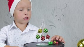 Cheerful child in a santa claus hat eats candy next to a small christmas tree
