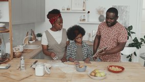 Medium slowmo shot of cheerful African American family of three cooking together at home watching video recipe on digital tablet making delicious apple pie