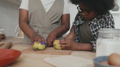 Tilting-up slowmo of African American mother and her little son cutting apples while cooking apple pie together standing by kitchen table at cozy home