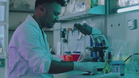 Handsome Ethnic Biochemist Working in a Modern Laboratory Wearing a White Lab Coat and Surgical Gloves. Lab Worker Looking at Bacteria Strains Through a Microscope Collecting Information.