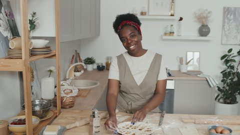 Medium slowmo portrait of smiling young African American woman posing for camera while making delicious apple pie standing by table in modern kitchen