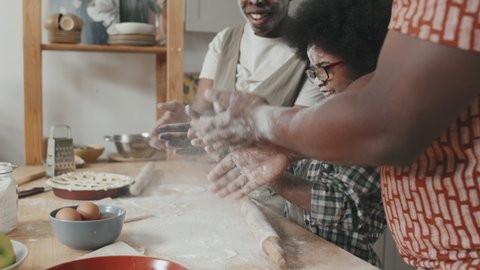 Slowmo shot of African-American family of three clapping hands with white flour while cooking homemade apple pie at kitchen table having fun together