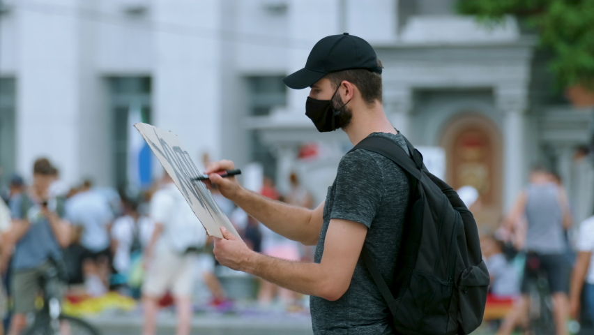 Protester man on rally in covid-19 mask draws sign poster. Rebel on city revolt, resistance strike. Picket activist drawing demonstration banner placard protesting. Royalty-Free Stock Footage #1077744692