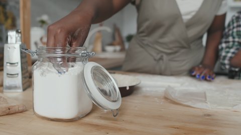 Slowmo close-up of female hand taking some flour from glass jar while little boy rolling out dough cooking together at home