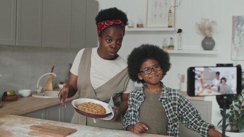Medium slowmo shot of African-American mother and son standing by kitchen table recording video recipe of homemade apple pie on smartphone