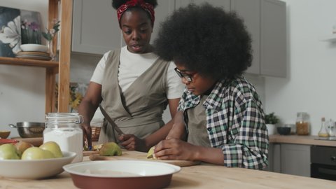 Slowmo shot of cute African American boy cutting fresh apples for apple pie and his mother helping him standing together by table at modern kitchen