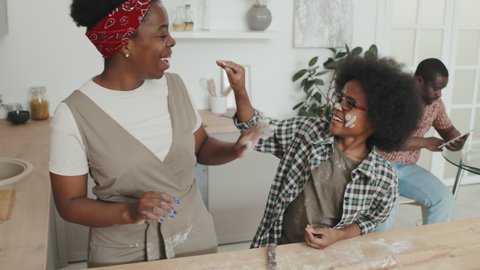 Medium slowmo shot of joyful African American mother and son having fun with flour while cooking homemade apple pie at kitchen table and father using digital tablet in background