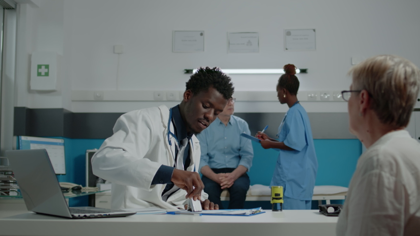Porfessional doctor using stamp on documents giving prescription to senior patient at desk in healthcare office. Medic advising elder woman about healing treatment and medicine