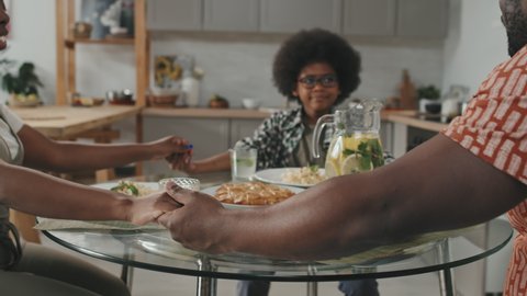 Slowmo shot of modern African American family of three sitting at dinner table with eyes closed holding hands and praying before eating